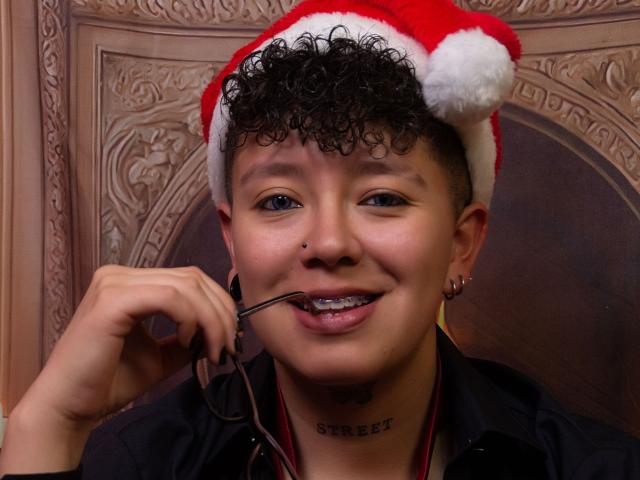 AllexNoa met krullend zwart haar, een rode kerstmuts op, een bril in zijn mond, lachend, een zwart shirt, een neusring en een "sweet" tattoo op zijn nek..