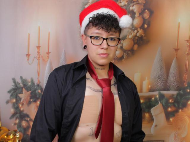 AllexNoa with curly black hair, glasses, and black shirt, wearing a red Santa hat and red tie, exposing a tattooed chest, and a flaccid, circumcised, tan-colored penis.