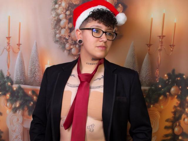 AllexNoa with curly brown hair, black glasses, and a red Santa hat, wearing a black blazer and red tie, exposing a tattooed chest, and large black ear plugs.