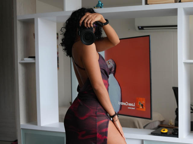 VivienneLaurent with curly black hair, wearing a tight, backless, red and purple dress, holding a camera to her face, showing her curvy ass and back in a white room.