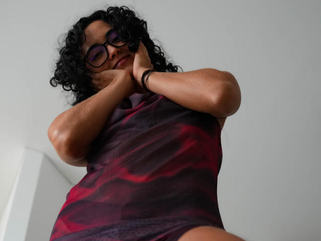 VivienneLaurent, dark curly hair, purple glasses, red and black tie-dye top, hands near face, looking down, low angle, wearing black wristbands, white background.