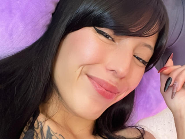 Close-up of AizenMyoo with black hair and bangs, smiling with eyes closed. She has fair skin, pink lipstick, and black winged eyeliner. Her right hand with black and red nails is near her face. She has a tattoo visible on her left collarbone. Purple background. Image has a soft, slightly blurry texture. AizenMyoo's relaxed, confident expression is central.