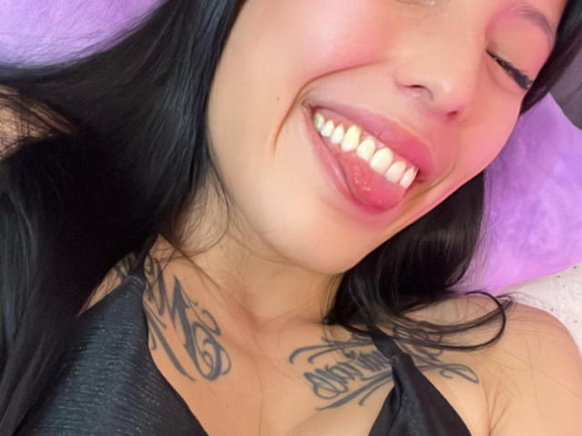 Close-up of AizenMyoo's face with black hair, fair skin, and pink blush on her cheeks. She's sticking out her tongue with white teeth visible. AizenMyoo has two tattoos on her chest: one in cursive script on the left and another with more intricate script on the right. She's wearing a black strap, and the background is a soft purple pillow. The image is candid, with natural lighting highlighting her teeth and tattoos. The composition focuses on her upper chest and face.