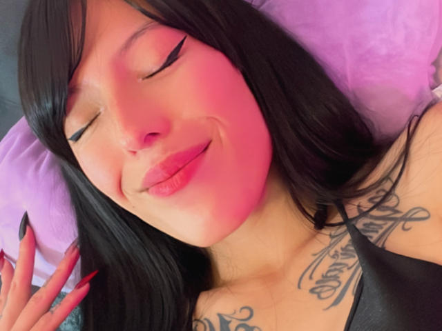 AizenMyoo with closed eyes, pink-lit face, black eyeliner, pink lipstick, black hair, smiling, visible tattoos on chest, black top, pink pillow, black nails.