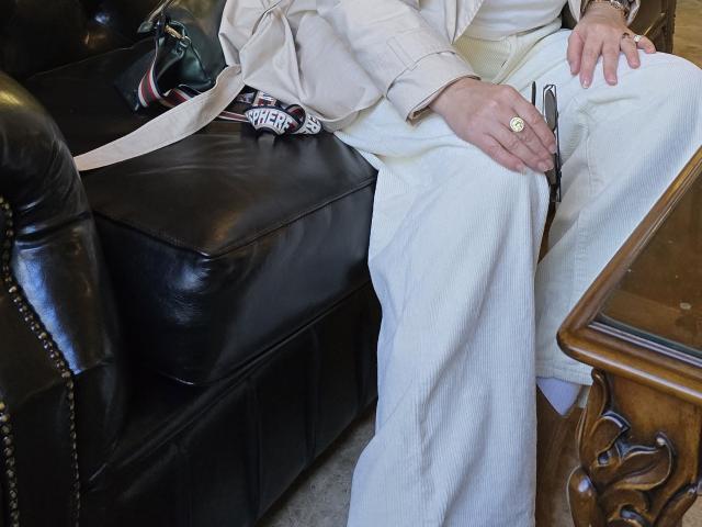 Los pantalones blancos de MonaCool, ligeramente holgados, con un bolsillo abotonado, sentada en un sofá de cuero negro, vistiendo una chaqueta beige, sosteniendo un bolígrafo y con un anillo de oro en el dedo..