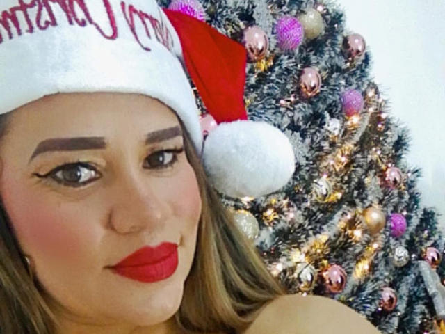 CharllotI with red lipstick, wearing a Santa hat, smiling at the camera, brown eyes, and blonde hair, in front of a decorated Christmas tree with pink and gold ornaments.