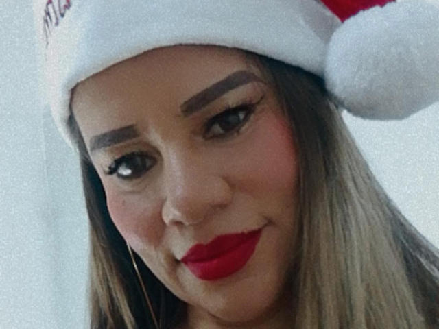 Close-up of CharllotI wearing a red and white Santa hat. CharllotI has light brown skin, thick brown eyebrows, long brown hair, and bright red lipstick. CharllotI's eyes are slightly downcast with long eyelashes. The Santa hat has a red ribbon and a white fluffy pom-pom. CharllotI's small gold hoop earrings are visible. The background is blurred and light-colored. The image has a soft, grainy texture.