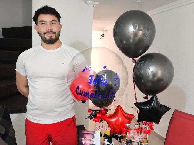 NiganDuquette, muscular with dark curly hair and beard, wears white tee and red shorts, holding gray balloons and clear balloon with "Cumpleaños" text, smiling.