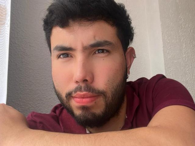 NiganDuquette has a dark curly haircut, a beard, and a maroon shirt. He's looking slightly confused with his arms crossed, showing a small nose piercing on his left ear.