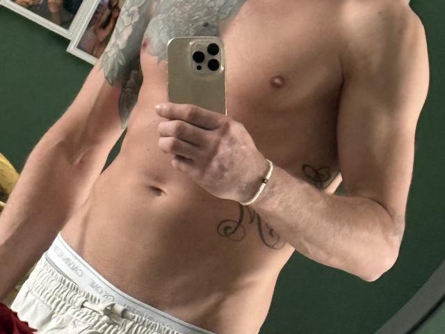 Mamba69's shirtless, muscular, white-skinned torso with tattoos, holding a phone, wearing white Calvin Klein underwear, visible chest and abs, and a wrist bracelet.
