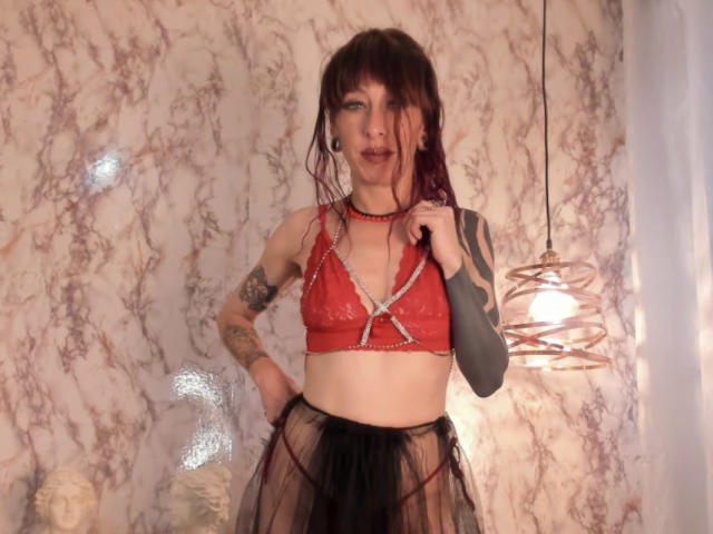 Grethel: Mulher com sutiã de renda vermelha, saia preta transparente, tatuagens e cabelo vermelho escuro, em pé, com a mão na cintura, olhando para a câmera, colar vermelho, parede branca, luminária pendente..