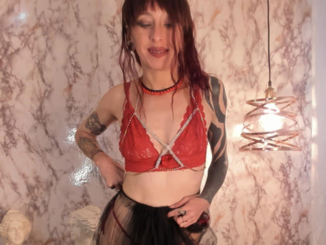 Grethel, una mujer con un top de encaje rojo, pequeños senos, un brazo tatuado, cabello rojo oscuro, una falda negra transparente, un collar de cuentas, una lámpara colgante, sonriendo, en una habitación con poca luz..