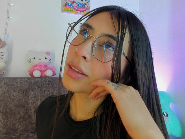 Macarenaa69 with long black hair, glasses, nose ring, and black shirt, looking at camera with hand on cheek, showing tattoo on wrist, and pink lips.