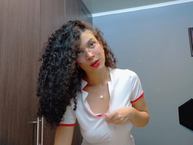 DylanyAllly69, avec ses cheveux noirs bouclés, son rouge à lèvres rouge, ses lunettes, et sa tenue de nurse blanche avec des détails rouges, tire sur le haut de sa tenue pour révéler de petites seins fermes, et porte un collier en or..