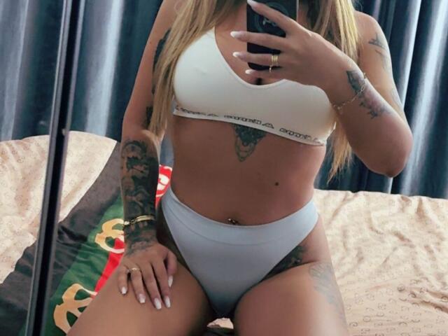 NicolSNaya mit gebräunter Haut, blonden Haaren, Tattoos an Armen und Bauch, einem weißen Sport-BH und hellblauen, hochgeschnittenen Unterhosen, macht ein Selfie vor einem Spiegel. Brüste, Hintern und Vulva sind sichtbar..