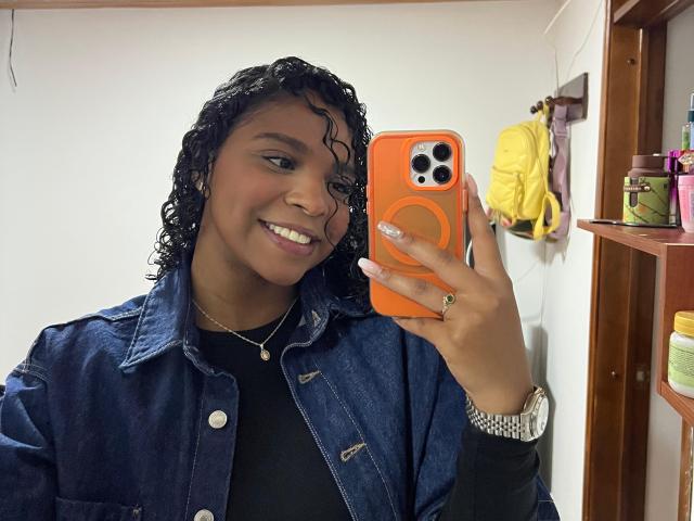 LisaMendez, met krullend zwart haar, lachend, draagt een denimjas, een zwarte top, houdt een oranje telefoon vast, en draagt een zilveren horloge, een ring, een ketting. Op de achtergrond is een gele rugzak te zien..