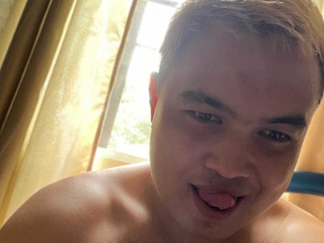 SexyJojo with short blonde hair, fair skin, and blue eyes, sticking out his tongue. He's shirtless, showing a smooth, bare chest. The image is a selfie.