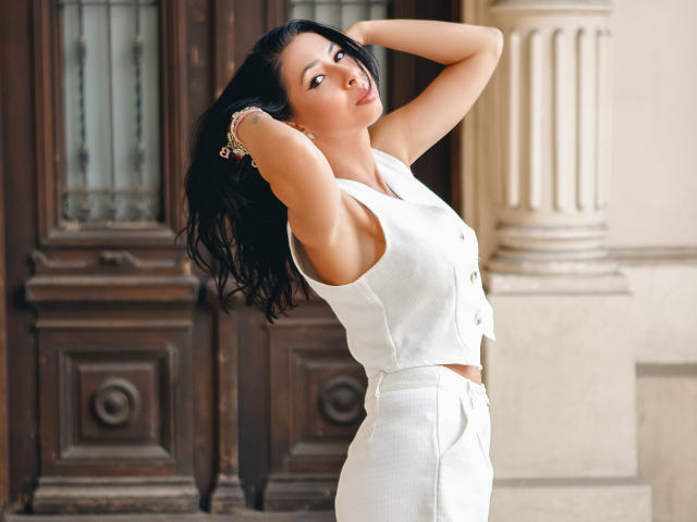 SarraRoses with arms raised, wearing a white sleeveless vest and matching pants, showing her tits and ass, standing in front of a wooden door.
