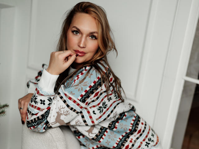 EvelynCute leans forward, resting her chin on her right hand. She has light brown, wavy hair and green eyes. She wears a colorful, patterned sweater with red, black, and blue geometric designs. Her left arm is draped over a white cushion. Her right hand touches her lips slightly. The image has a soft, natural light. Her expression is slightly curious and relaxed. The background is a white, paneled wall. The composition is centered with EvelynCute's face in the upper right and her body in the lower left. The texture of her sweater is slightly fuzzy.