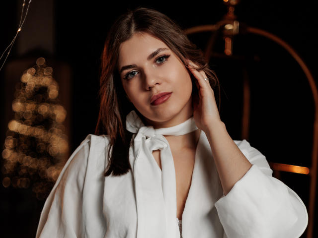 BelleLovely with wavy brown hair, wearing a white robe, hand on her face, looking at the camera, soft smile, light makeup, ring on her right hand, bokeh Christmas lights.