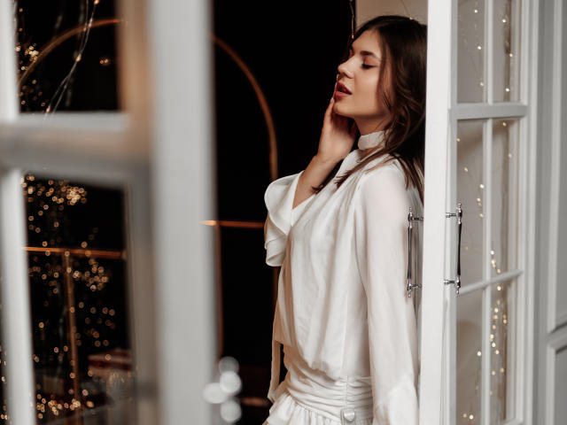 BelleLovely stands by an open window, eyes closed, wearing a white silky robe, hand on her neck, brown wavy hair, soft bokeh lights in the background.