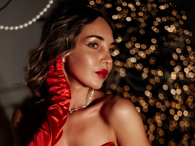 Close-up of LettieSweet with wavy brown hair, wearing a red satin fabric draped over her shoulder and a pearl necklace. She has red lipstick and a slightly parted, sultry expression. Bokeh lights in the background create a glowing effect. Her skin is smooth and she has a light tan. The red fabric and lights contrast with her skin tone. The lighting highlights her cheekbones and upper chest. The overall composition is dramatic and elegant.
