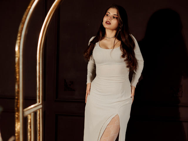 AdeliaLove with long brown hair, white ribbed dress, cleavage, hand in pocket, white lace thigh slit, standing, shadow on wall, gold curtain pole, dark background.