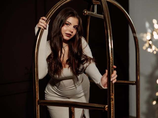 AdeliaLove with long brown hair, wearing a tight, beige, ribbed dress, holding a brass luggage cart, smiling seductively, showing her boobs slightly, in a dimly lit room.