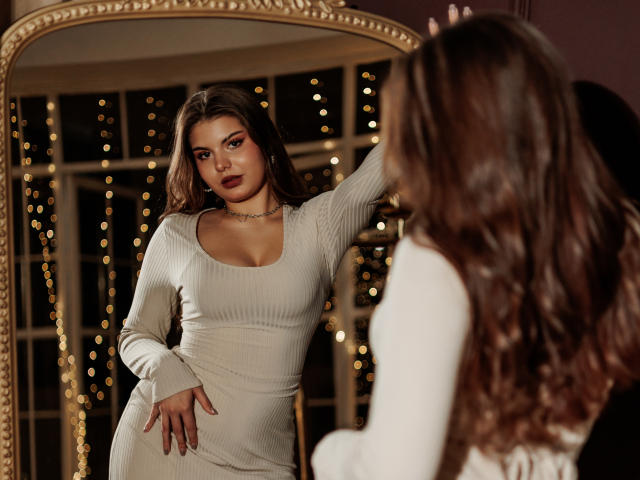 AdeliaLove stands in front of a mirror, wearing a tight, beige, rib-knit dress with a deep scoop neckline. She has long, wavy brown hair and dark makeup, including red eyeshadow and lipstick. AdeliaLove's right arm is raised, touching her hair, while her left hand rests on her dress. The reflection in the ornate, gold-framed mirror is visible. The image has warm, soft lighting, emphasizing her figure and the texture of her dress. Her expression is confident and slightly sultry. The background features blurred, glowing lights.