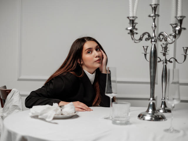 MilanaKitty with long brown hair, black suit, and white shirt, resting her head on her hand, looking slightly to the side, sitting at a white table with a candelabrum.