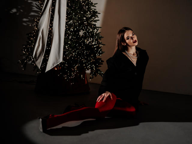 SophieKitty knielend voor een kerstboom, met rode leggings, een zwarte jas en donkere lipstick, verleidelijk naar de camera kijkend, met een schaduw op de vloer..