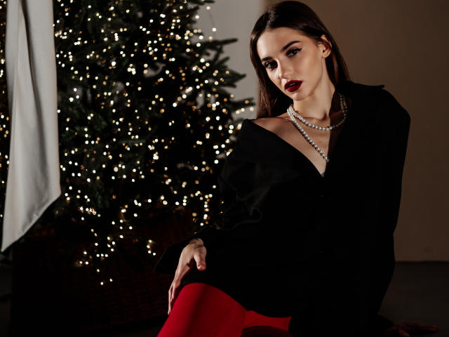 SophieKitty met donkerrode lipstick, een zwarte top met open schouders, een parelketting en rode leggings, zittend naast een kerstboom met lichtgevende lampjes. Haar borsten zijn gedeeltelijk zichtbaar..