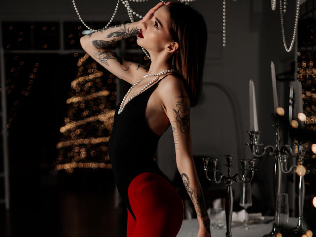 SophieKitty met tatoeages, gekleed in een zwarte top en rode broek, poseert met haar hand op haar hoofd, draagt een parelketting, staat bij kaarsen, kijkt omhoog, donker haar, rode lipstick..