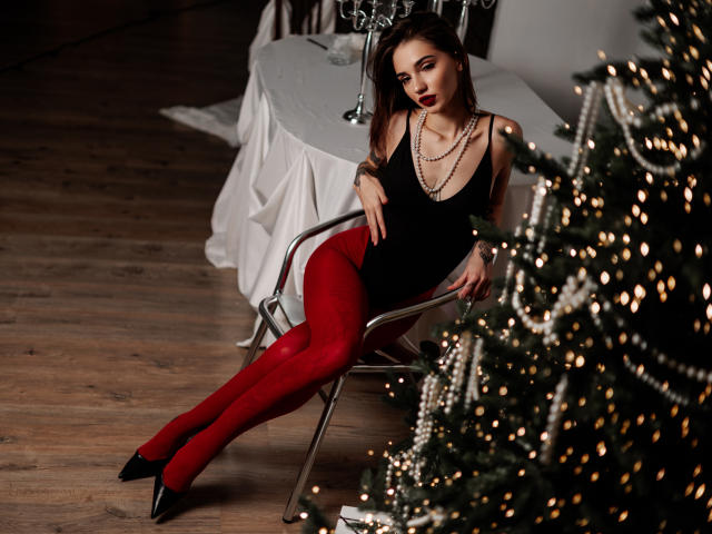 SophieKitty zit op een metalen stoel, gekleed in een zwart tanktop, rode leggings, zwarte hakken en een ketting. Ze staat naast een kerstboom met lichtjes..