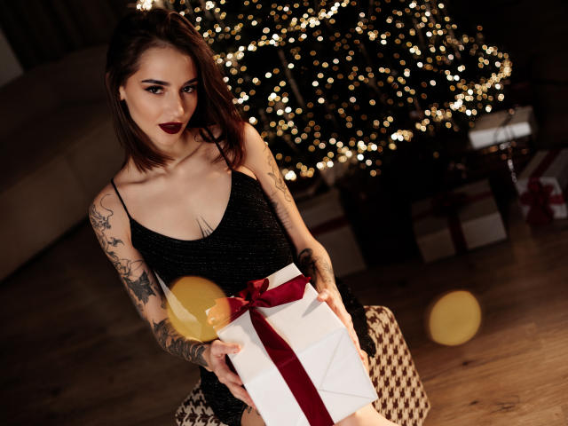 SophieKitty met tatoeages, donkere lipstick, een zwarte glinsterende jurk, die een cadeau met een rode strik vasthoudt, met een gele ballon, staand voor een kerstboom met bokeh-lichtjes..