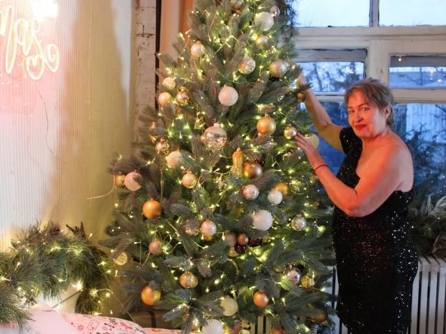OlgaLuck, una donna corpulenta con i capelli biondi corti, in un abito nero ricoperto di paillettes, sta decorando un albero di Natale con ornamenti dorati e argentati, toccando un ramo..