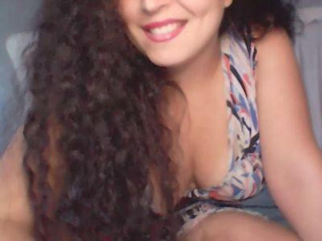 A close-up photo of SexCoppia6 with long, curly dark hair cascading over her right shoulder. She has a warm, smiling face with red lipstick. She's wearing a low-cut, floral-patterned dress that reveals significant cleavage. Her left arm is visible, and her right knee is slightly bent. The image is slightly blurry. The focus is on her face and chest. She has a light brown skin tone. The overall composition is casual and intimate.