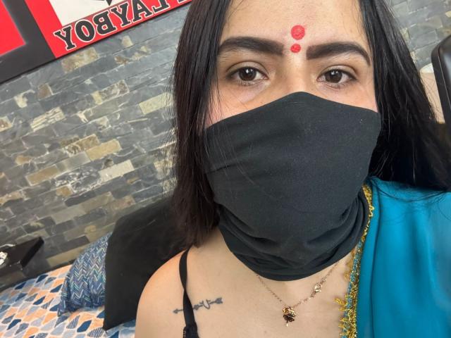 Dalia Dubey mit schwarzer Maske, roten Punkten auf der Stirn, schwarzem Haar, braunen Augen, Halskette, Tattoo auf der Schulter, blauem und goldenem Gewand, Steinmauer im Hintergrund, sichtbares Bett..
