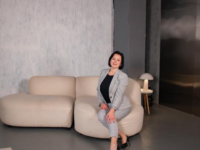 MarieSho with short black hair, gray suit, black top, sitting on a beige sofa, smiling, hands on her knees, gray wall behind, small lamp on right, black heels.