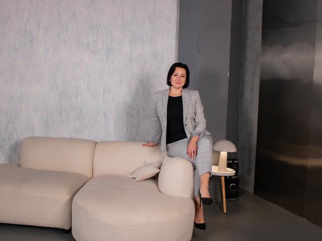 MarieSho with short black hair, wearing a gray plaid suit, black top, and black heels, sitting on a beige sofa, her hand on the cushion, legs crossed.