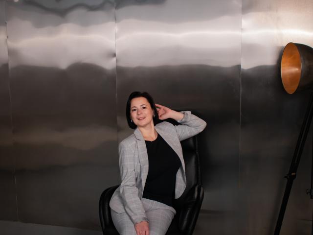 MarieSho, with short brown hair, wearing a grey plaid jacket and black top, sits in a black chair, hand in hair, smiling, next to a standing lamp.