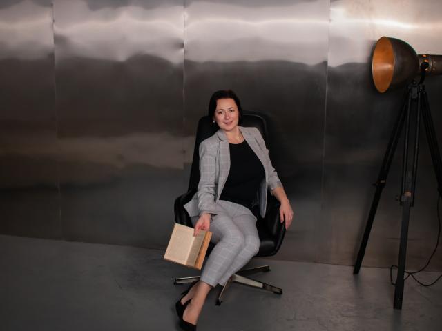 MarieSho, with small tits, sits in a black chair, wearing a gray jacket and black pants, holding a book, against a metallic wall, with a tripod lamp to the right.