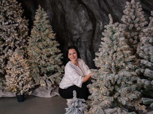 MarieSho with medium tits, dark hair, smiling, wearing white shawl, black pants, squatting between frosted Christmas trees, glowing white lights, rock cave background, daytime.