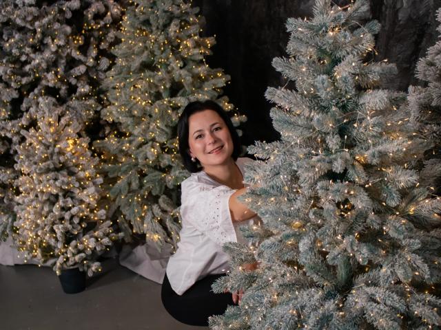 MarieSho with tits visible, smiling, wearing a white sleeveless top, black pants, crouching between two frosty Christmas trees with warm yellow lights, in a dark, cozy setting.