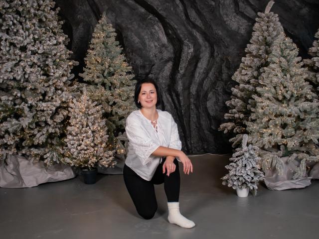 MarieSho, with medium tits, black hair, white blouse, black pants, white socks, kneeling in front of snow-covered Christmas trees, smiling. She's cute, with a relaxed, happy expression.