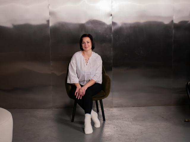 MarieSho sits on a green chair, wearing a white lace-up shirt, black pants, and white socks, smiling with hands on her knees, against a metallic wall.