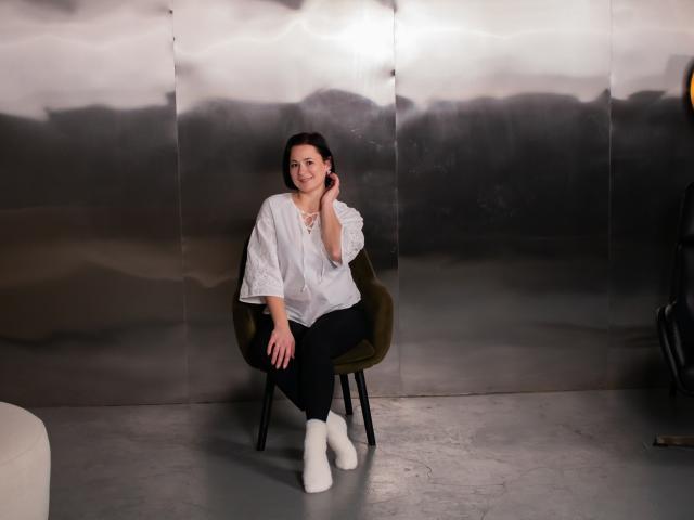 MarieSho with dark hair, white blouse, black pants, and white boots, sitting in a green velvet chair, smiling, hand on cheek, against a metallic wall.