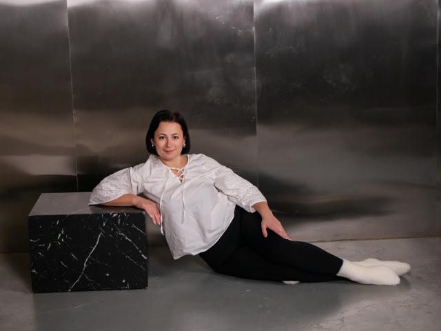 MarieSho with tits covered by a white shirt, black pants, and white socks, laying on a marble block, against a metallic wall, smiling at the camera.