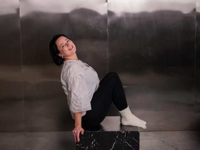 MarieSho with short black hair, smiling, leaning back on a marble block, wearing a gray shirt, black pants, and white socks, against a metallic wall.