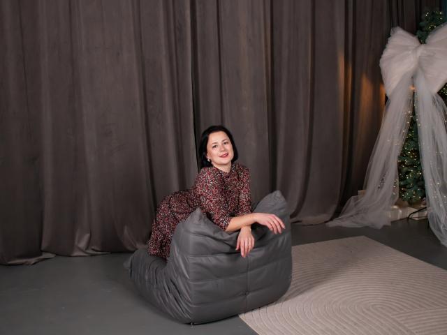MarieSho with medium tits, red floral dress, black curly hair, smiling, leaning on gray leather beanbag, standing on white rug, next to Christmas tree with big white bow.